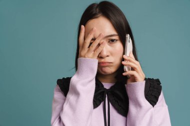 Genç Asyalı kadın yüzünü örtüyor ve cep telefonuyla konuşuyor. Mavi arka planda izole edilmiş.