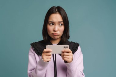 Genç Asyalı kadın mavi arka planda izole edilmiş bir şekilde cep telefonuyla çevrimiçi oyun oynuyor