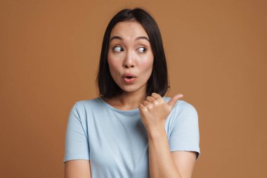 Genç Asyalı kadın şaşkınlığını dile getiriyor ve bej arka planda izole edilmiş bir şekilde işaret ediyor.