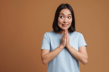Kafası karışmış Asyalı kadın avuçlarını bir arada tutarken yana bakıyor bej arka planda izole edilmiş.