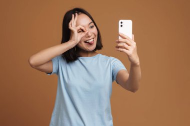 Genç Asyalı kadın bej arka planda izole edilmiş cep telefonuyla selfie çekerken el kol hareketi yapıyor.