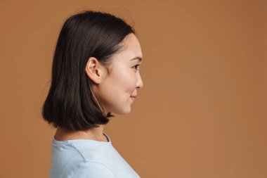 Bej arka planda izole edilmiş, profilde poz verirken gülümseyen Asyalı genç bir kadın.