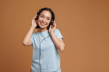 Bej arka planda izole edilmiş müzik dinlerken gülümseyen kulaklıklı genç Asyalı kadın.