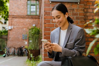 Kulaklıklı Asyalı kadın bina kenarında otururken cep telefonu kullanıyor.