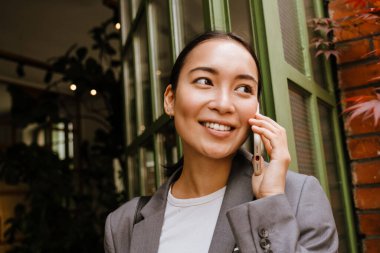 Asyalı genç iş kadını sokakta ayakta dururken gülümsüyor ve cep telefonuyla konuşuyor.