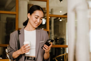 Kafede durup kahve içen ve cep telefonu kullanan Asyalı genç iş kadını.