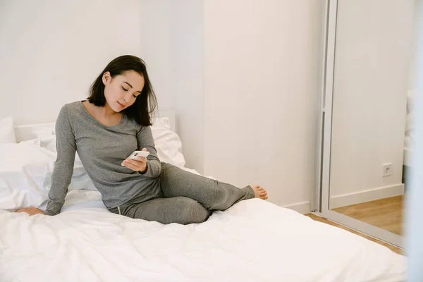 Stock image Young asian girl in home suit with phone sitting on the bed at home