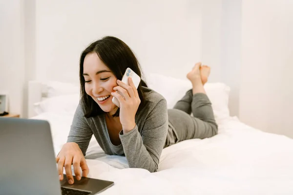 身穿家居服 面带微笑的年轻亚洲女孩躺在床上 拿着笔记本电脑 在家里打电话 — 图库照片