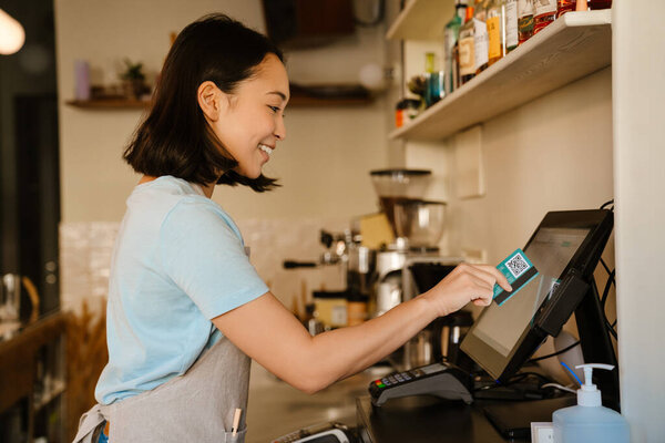 Asian barista woman wearing apron smiling while working with cash register in cafe