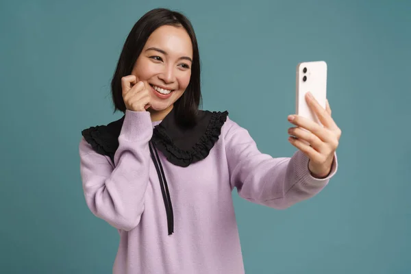 年轻的亚洲女人一边笑着一边用蓝色背景的手机自拍 — 图库照片