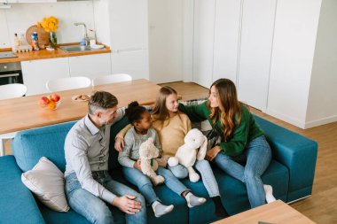 Mutlu bir aile kanepede oturur, evlâtlık kız kardeşler evdeki geniş oturma odasında oyuncaklarla oynarlar. Evlat edinme kavramı.