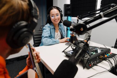 Genç beyaz adam gitar çalarken mutlu kadın radyo stüdyosunda dizüstü bilgisayarda podcast yapıyor.