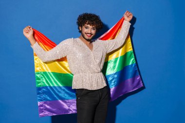 Mavi arka planda izole edilmiş kameraya gülümseyen gökkuşağı bayraklı genç kıvırcık adam.