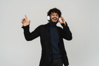 Gri arka planda kulaklıkla dans eden genç ve mutlu Hintli bir adam.