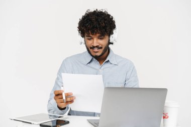 Genç Hintli ofis çalışanı dizüstü bilgisayarla oturmuş kağıda bakıyor ve gri arka planda bir kalem tutuyor.