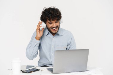 Genç Hintli gülümseyen ofis çalışanı dizüstü bilgisayarla oturmuş gri arka plan üzerinden kulaklıkla ekrana bakıyor.