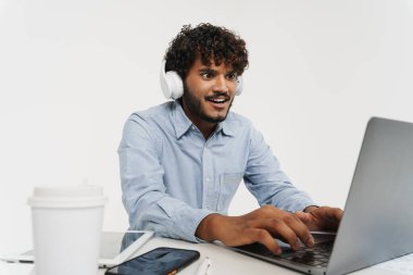 Yetişkin Hintli kıvırcık ofis çalışanı dizüstü bilgisayarın önünde oturuyor ve gri izole arka plandan ekrana bakıyor.