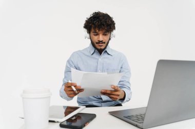 Genç Hintli kıvırcık ofis çalışanı, dizüstü bilgisayarı kulaklıkla oturuyor ve izole edilmiş gri arkaplandaki belgelere bakıyor.