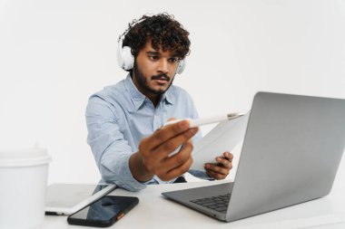 Genç Hintli ciddi ofis çalışanı dizüstü bilgisayarı ve ekranda kalemle gösterilen belgelerle oturuyor.