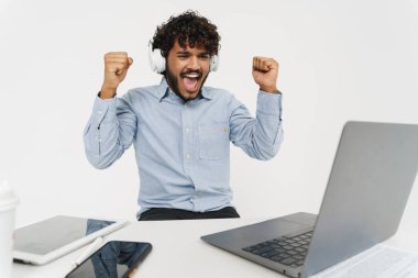 Genç Hintli mutlu ofis çalışanı dizüstü bilgisayarın önünde oturuyor ve gri izole arka planda kollarını kaldırarak kutluyor.