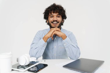 Yetişkin Hintli ofis çalışanı, ofis masasının yanında oturuyor ve elleri katlanmış, gri izole arka planda oturuyor.