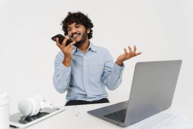 Yetişkin Hintli ofis çalışanı, Gri izole arka planda dizüstü bilgisayarın önünde telefonla konuşuyor.