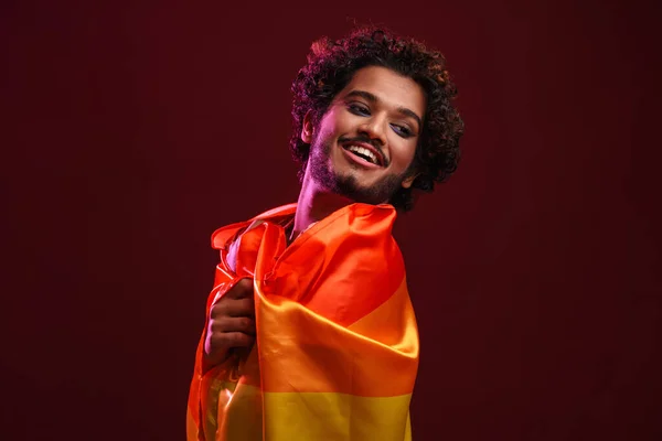 年轻的卷曲男子裹着彩虹旗 微笑着看着被红色背景隔开的相机 — 图库照片