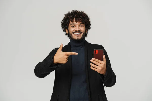 若い幸せな笑顔インド人男性携帯電話を保持し それを指して 孤立した灰色の背景の上にカメラを見て — ストック写真