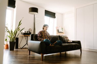 Beyaz adam evdeki kanepede dinlenirken dizüstü bilgisayar kullanıyor.