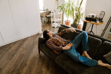 Gözlüklü beyaz adam evdeki kanepede dinlenirken cep telefonu kullanıyor.