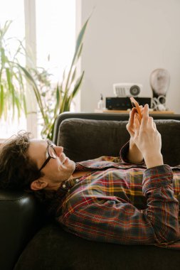 Beyaz adam evde kanepede dinlenirken gülümsüyor ve cep telefonu kullanıyor.