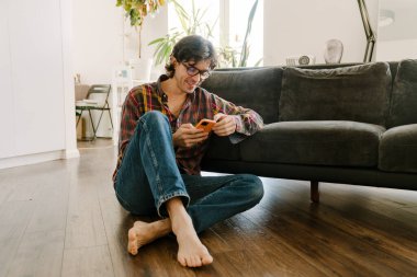 Gözlüklü beyaz adam evde otururken cep telefonu kullanıyor.