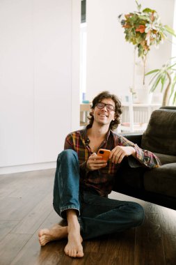 Gözlüklü beyaz adam evde otururken cep telefonu kullanıyor.