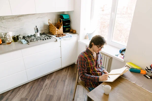 Bílý Muž Nosí Brýle Píše Poznámky Zatímco Sedí Doma Kuchyni — Stock fotografie