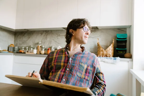 Bílý Muž Nosí Brýle Píše Poznámky Zatímco Sedí Doma Kuchyni — Stock fotografie