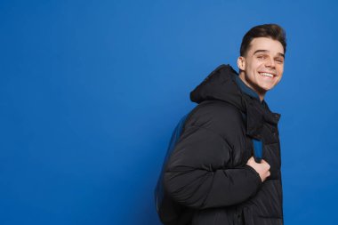 Sırt çantalı genç beyaz adam gülümsüyor ve mavi arka planda izole edilmiş kameraya bakıyor.