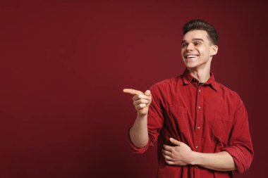 Gömlek giyen genç beyaz bir adam kırmızı arka planda yalnız bir şekilde parmak gösterip gülüyor.