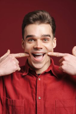 Gömlek giyen genç beyaz adam kırmızı arka planda izole edilmiş gülüşünü işaret ediyor.