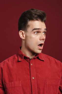 Kırmızı arka planda izole edilmiş kameraya şaşırdığını ifade eden tişört giyen genç beyaz bir adam.
