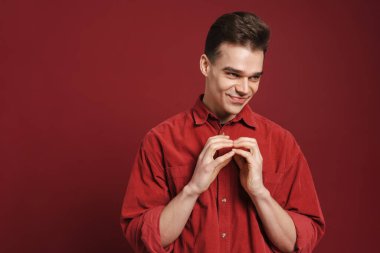 Genç beyaz adam parmaklarını birarada tutuyor ve kırmızı arka planda izole bir şekilde gülümsüyor.