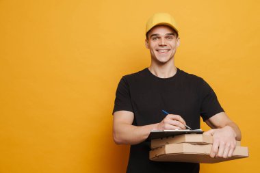 Sarı arka planda izole edilmiş pano ve pizza kutularıyla poz verirken gülümseyen genç teslimatçı.