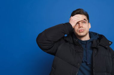 Alnını tutup yukarı bakan yorgun beyaz adam mavi arka planda izole edilmiş.