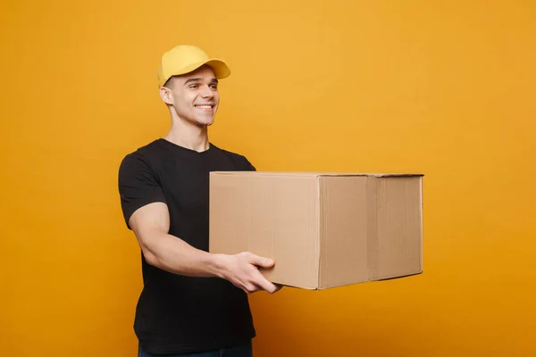 Sarı Arka Planda Karton Kutuyla Poz Verirken Gülümseyen Genç Teslimatçı — Stok fotoğraf