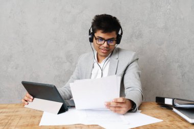 Genç mutlu Hintli işadamı ofiste çalışırken tablet bilgisayar ve kulaklık kullanıyor.