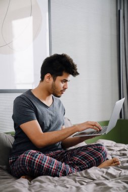 Genç, yakışıklı Hintli bir adam Lotus 'ta yatağında oturmuş dizüstü bilgisayara poz veriyor ve evdeki rahat yatak odasında daktilo yazıyor.