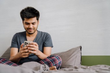 Genç Hintli gülümseyen adam telefonunu kaydırıyor evinde yatağında lotus pozu veriyor.
