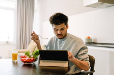 Genç Hintli adam tablet üzerinde kayıyor ve evde bir bardak portakal suyu ile taze salata yiyor.