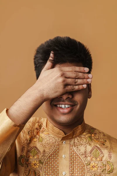 Young brunette indian man smiling and covering his eyes with hand isolated over yellow background