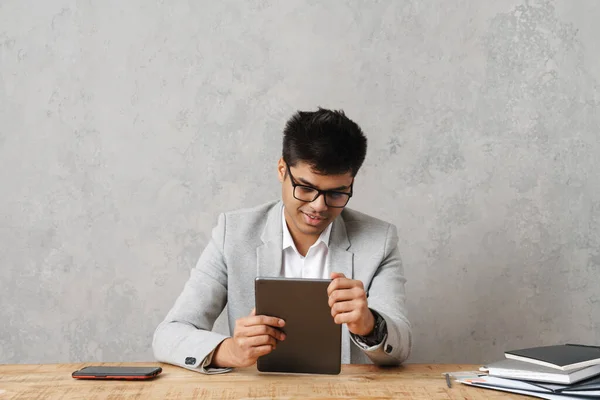 若いブルネットのインド人ビジネスマンがタブレットコンピュータを使用してグレーの壁の上にオフィスのテーブルに座って — ストック写真