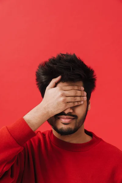 Young brunette indian man smiling and covering his eyes isolated over red background